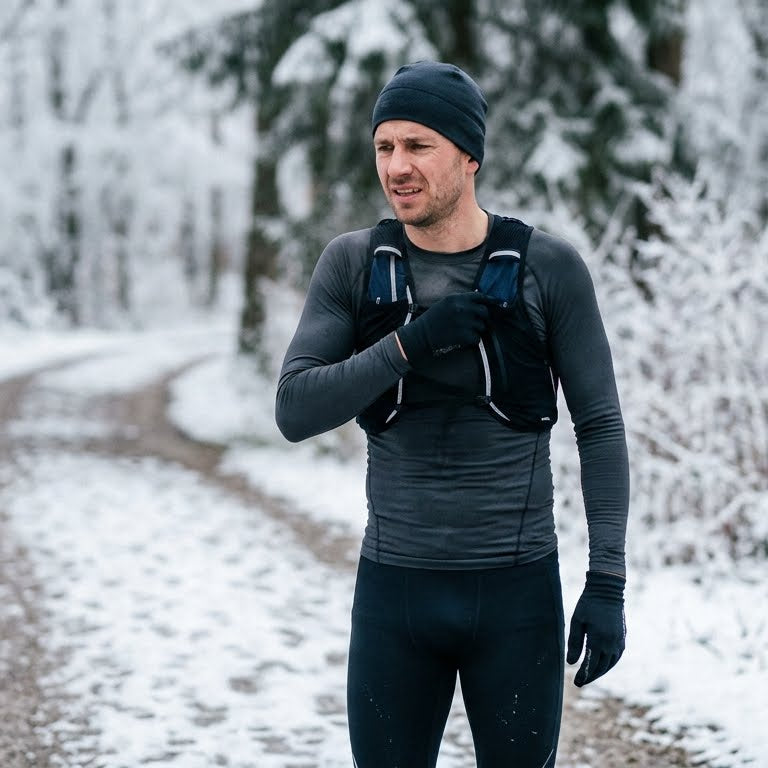 A male runner in cold weather gear adjusting his tight synthetic base layer on a snowy trail, demonstrating the discomfort and friction caused by winter running layers.