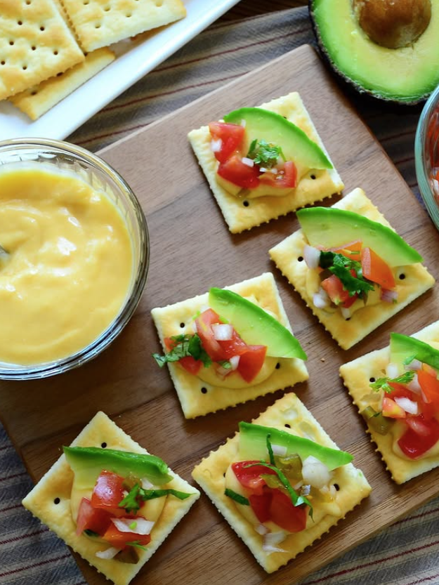 Crackers with avocado and tomato on a wooden board with cheese and avocado in the background