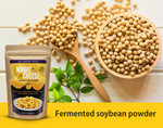 A product image of Living Jin Magic Cheese Cheddar Style Dairy-Free Powder in a brown kraft stand-up pouch. The pouch is displayed alongside a wooden bowl and spoon filled with whole soybeans, emphasizing the product's primary ingredient. A bold yellow banner at the bottom contains the text "Fermented soybean powder," highlighting the natural, plant-based origin of the vegan cheese alternative.